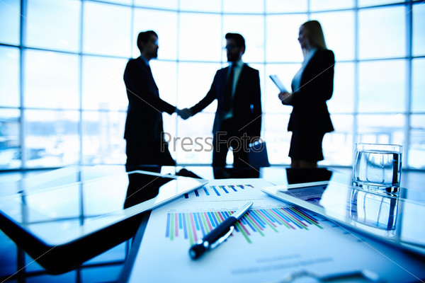 Technological devices, financial document with pen, glass of water at workplace on background of three business partners striking deal, stock photo