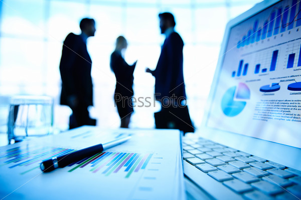 Laptop and financial document with pen at workplace on background of three business partners interacting