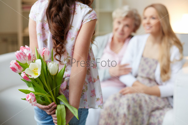 Rear view of little girl holding bunch of flowers behind back with her mother and grandmother on background