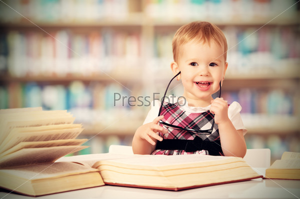 Happy funny baby girl in glasses reading a book in a library, stock photo