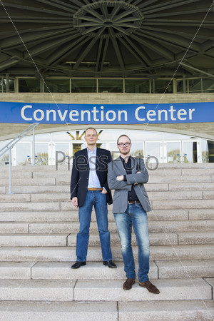 Two attendees of a congress at a trade fair posing on the steps leading to the front entrance of the convention center.