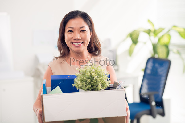 Portrait of Asian woman holding box with documents, plant and office objects and looking at camera with smile