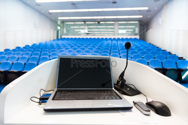 Laptop and microphone on the rostrum in empty conference hall with blue velvet chairs.