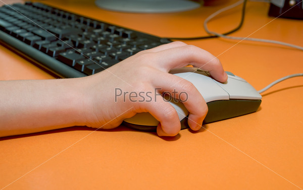 Child\'s right hand on the mouse on the background of computer keyboard on the orange table