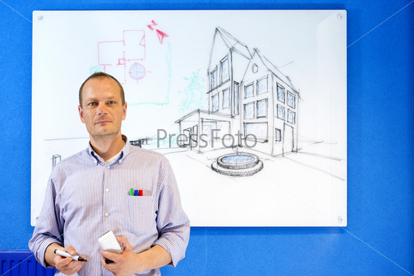 Architect, holding a white board marker, standing in front of a design sketch of a residential structure on the glass board behind him