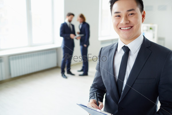 Young successful manager looking at camera while his colleagues talking on the background