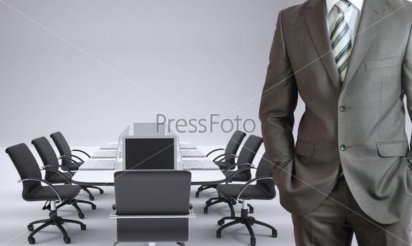 Businessman standing with hands in pockets. Conference table, chairs and laptops as backdrop
