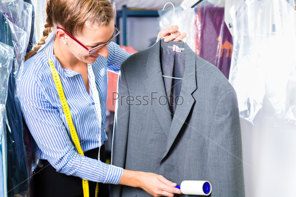 Female cleaner in laundry shop checking clean clothes removing lint with roller