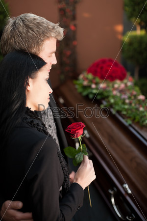 Morning man and woman on funeral with red rose standing at casket or coffin