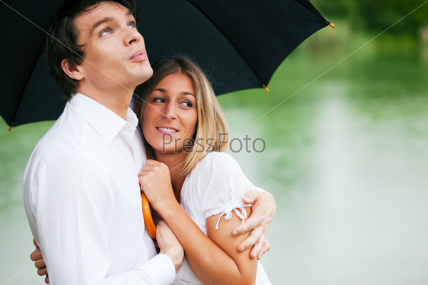 Couple (man and woman) at a lake in the rain with an umbrella, he is sheltering her from the drops, holding his girl in his arms
