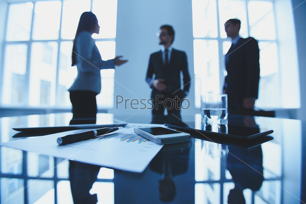 Image of business document and electronic devices at workplace with group of business people talking on background