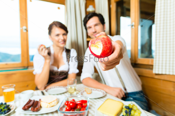 Couple in a traditional mountain hut having a meal, breakfasting with fruits, cold cuts, cheese and bread