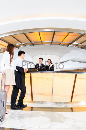 Asian Chinese woman and man arriving at front desk or reception of luxury hotel in business clothes with trolley