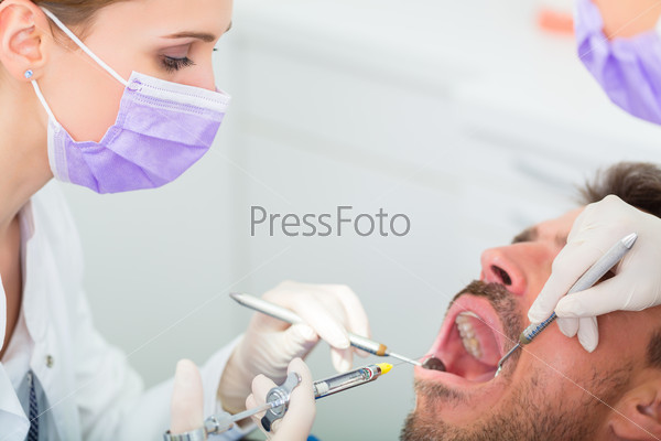Male patient with dentist and assistant in a dental\
treatment, wearing masks and gloves, giving anesthetization\
syringe