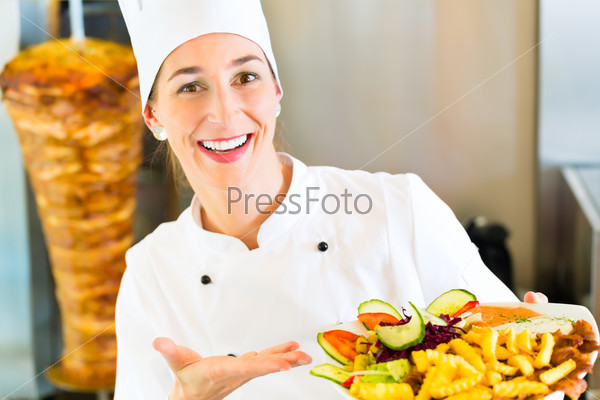 Doner kebab - friendly female vendor in a Turkish fast food eatery, holding plate with fries and kebab in front of skewer