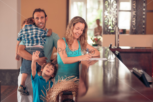 Middle-aged woman checking in, while her husband carrying a tired child, and her other son waving at the camera