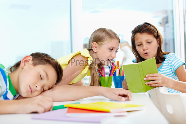 Cute schoolgirl showing her classmate her notes, tired boy napping on desk near by