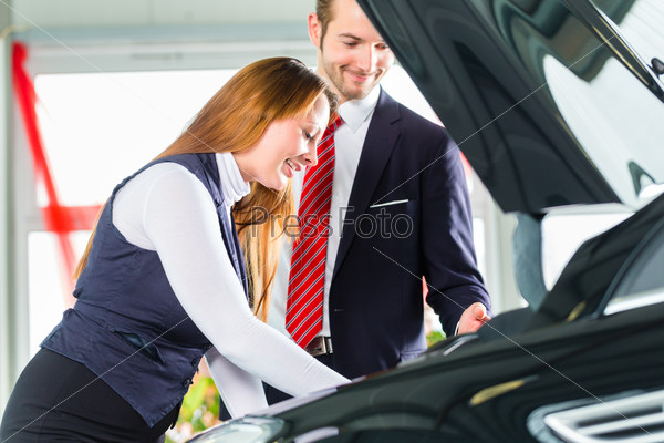 Seller or car salesman and female client or customer in car dealership presenting the engine performance of new and used cars in the showroom, the woman looks under the hood