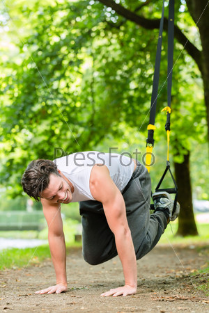 Young man exercising with suspension trainer sling in City Park under summer trees for sport fitness, stock photo