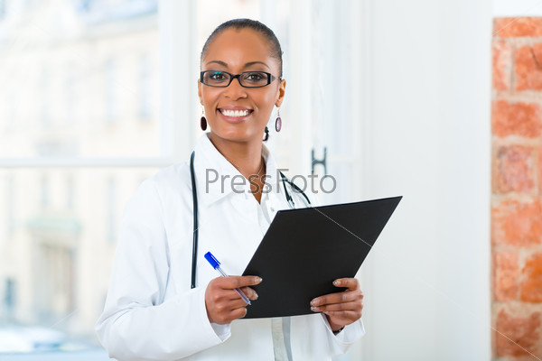 Young female black doctor standing at a window in clinic with a file or dossier