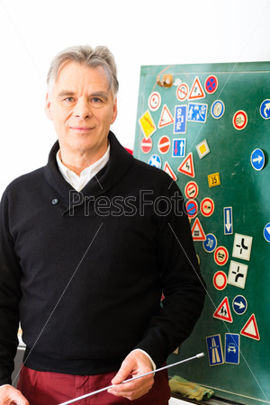 Driving school - driving instructor in his class, he standing in front of a blackboard and looking in the camera, in the background are traffic signs