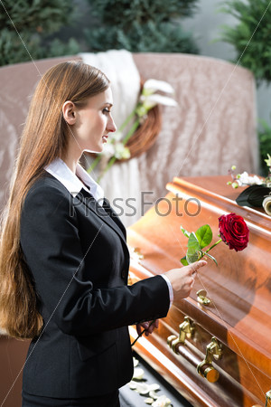 Mourning woman on funeral with red rose standing at casket or coffin