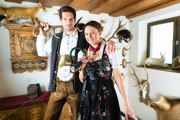 Young hunter with his wife and a bugle in front of a Wall with different horns, antlers and trophies in an alpine cabin, stock photo