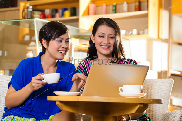 Asian female friends enjoying her leisure time in a cafe, drinking coffee or cappuccino and working on a laptop computer