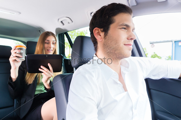 Young taxi driver driving businesswoman in taxi, she holding cup of coffee and a tablet computer, stock photo