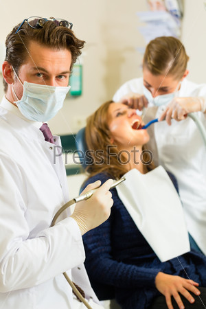 Dentists in his surgery holds a drill and looking at the viewer, in the background his assistant is giving a female patient a treatment