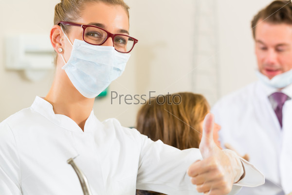 Dentists in his surgery holds a drill and looking at the viewer, in the background her colleague is giving a female patient a treatment, stock photo