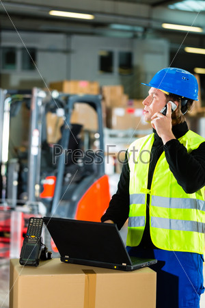 Warehouseman with protective vest, scanner and laptop in warehouse at freight forwarding company using a mobile phone