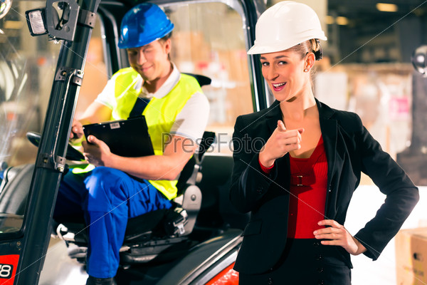 Forklift driver with clipboard at warehouse of freight forwarding company, female super visor or dispatcher pointing to the viewer