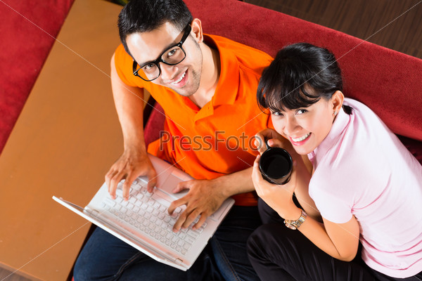 Индонезийская пара сидит на диване с ноутбуком, используя интернет для электронной почты и онлайн магазинов