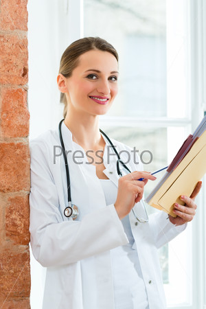 Young female doctor standing at a window in clinic writing in a file or dossier