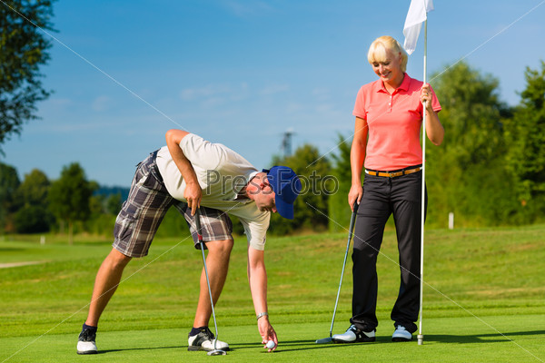 Young sportive couple playing golf on a golf course, he takes the ball from the hole