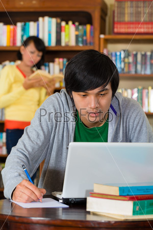Student - Young Asian man in library with laptop learning, a female student standing in the Background on a shelf reading book