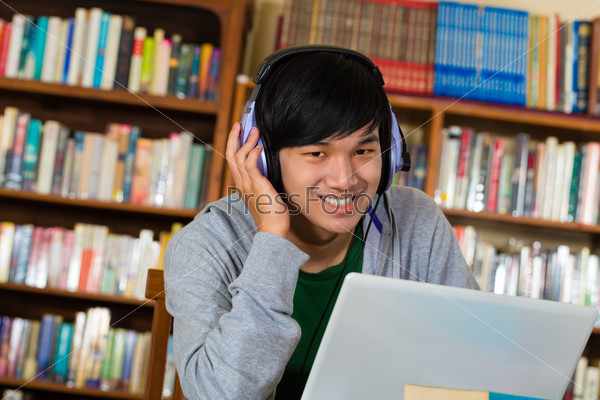 Student - Young Asian man in library with laptop and headphones learning, stock photo