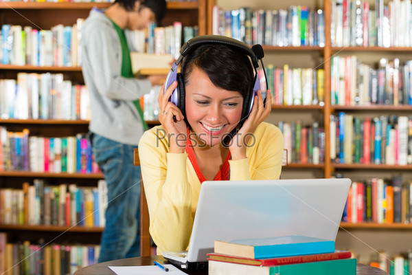 Student - Young woman in library with laptop and headphones\
learning, a male student standing in the Background reads a\
book