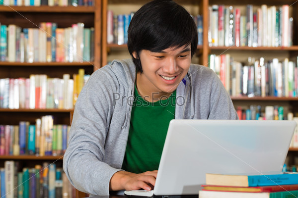 Student - Young Asian man in library with laptop and a pile of books learns