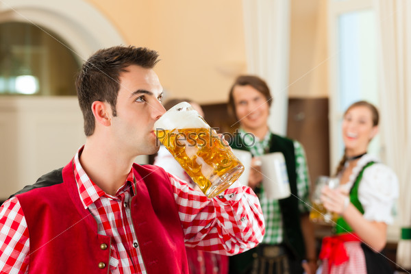 Young people in traditional Bavarian Tracht in restaurant or pub, one man is standing with beer stein in front, the group in the background