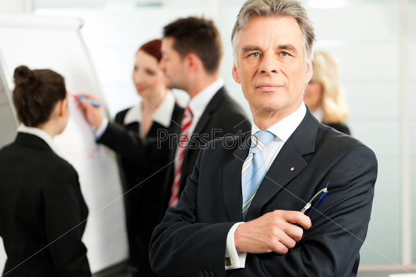 Business - team in an office, a colleague is standing in front of the flipchart, one female colleague is looking in to the camera