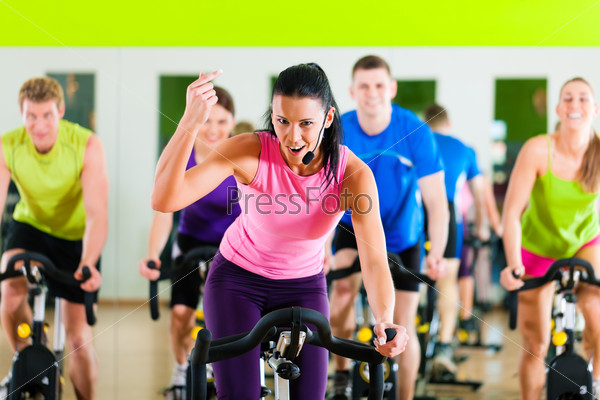 Group of five people - men and women - in gym or fitness club exercising their legs doing cardio training; the trainer is in front