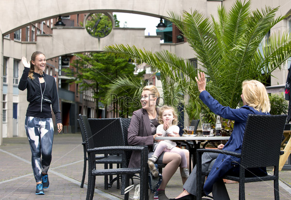 A group of three, very diverse women, meeting on a terrace. A sporty jogging woman waves at a senior lady, with a mother and her young daughter also sitting at the table in an urban setting.