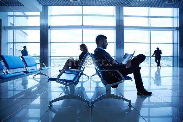 Two business partners reading at the airport on background of their colleagues by the window
