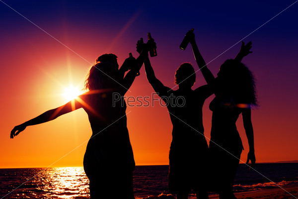 People (two couples) on the beach having party, drinking and having a lot of fun in the sunset (only silhouette of people to be seen, people having bottles in their hands with the sun shining through)