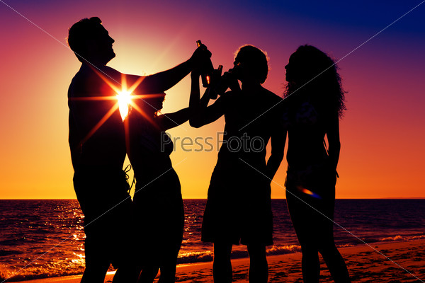 People (two couples) on the beach having a party, drinking and having a lot of fun in the sunset (only silhouette to be seen, people having bottles in their hands with the sun shining through)
