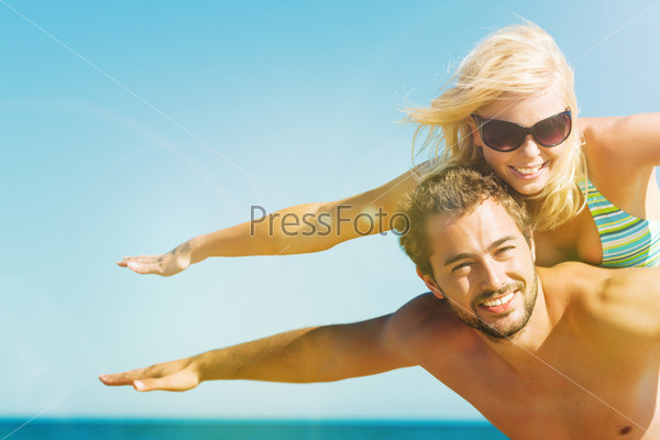Couple in love on beach in vacation - man carrying his wife on the back close to ocean, they are feeling visibly free and happy