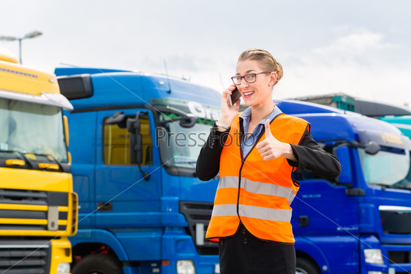 Logistics - female forwarder or supervisor with mobile phone, in front of trucks and trailers, on transshipment point