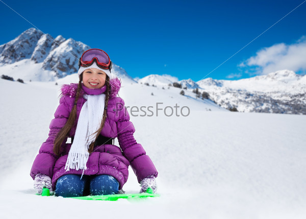 Happy girl sitting on sled with her hands lifted, wearing ski mask and purple coat, in the mountains, stock photo